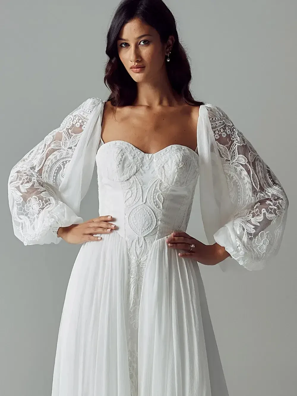 Woman in elegant white wedding dress with lace sleeves and fitted bodice stands confidently. Her hands rest on her waist, exuding grace and poise.
