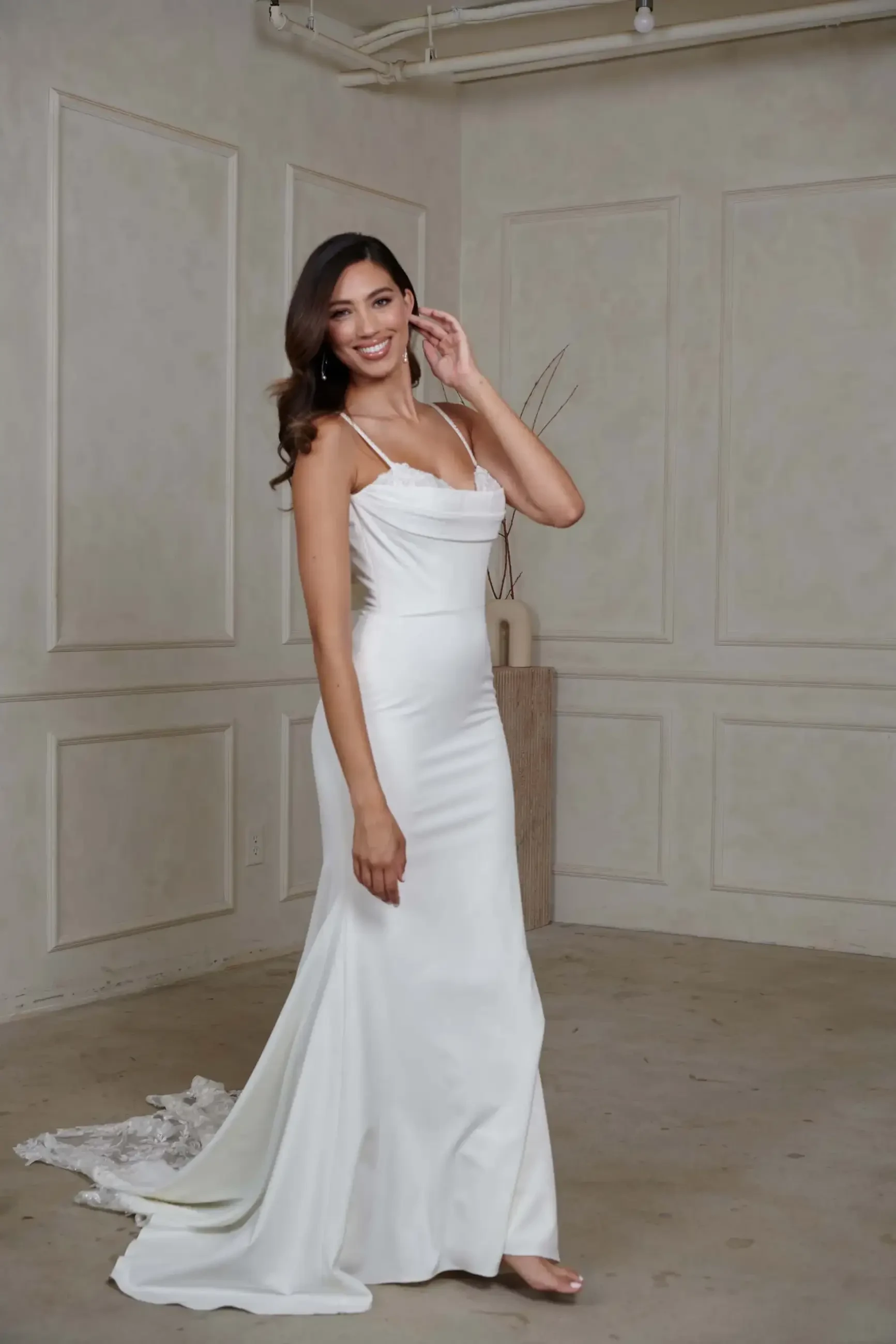 A woman in a sleek white gown with spaghetti straps stands barefoot in a neutral-toned room, smiling and touching her long wavy hair. Elegant and joyful.