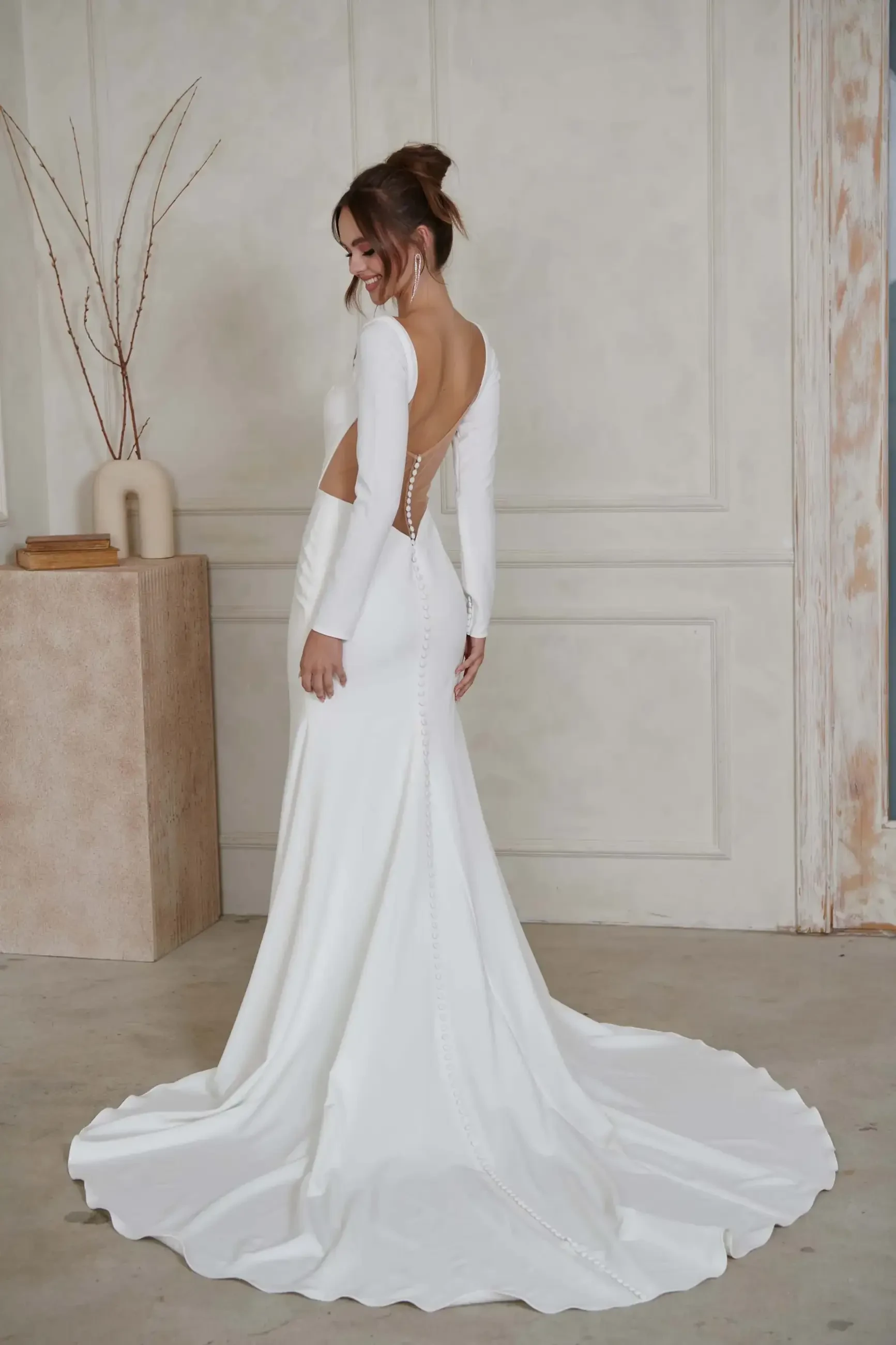 Woman in an elegant white wedding dress with a long train, standing in a minimalist room. The dress features an open back and button detailing.