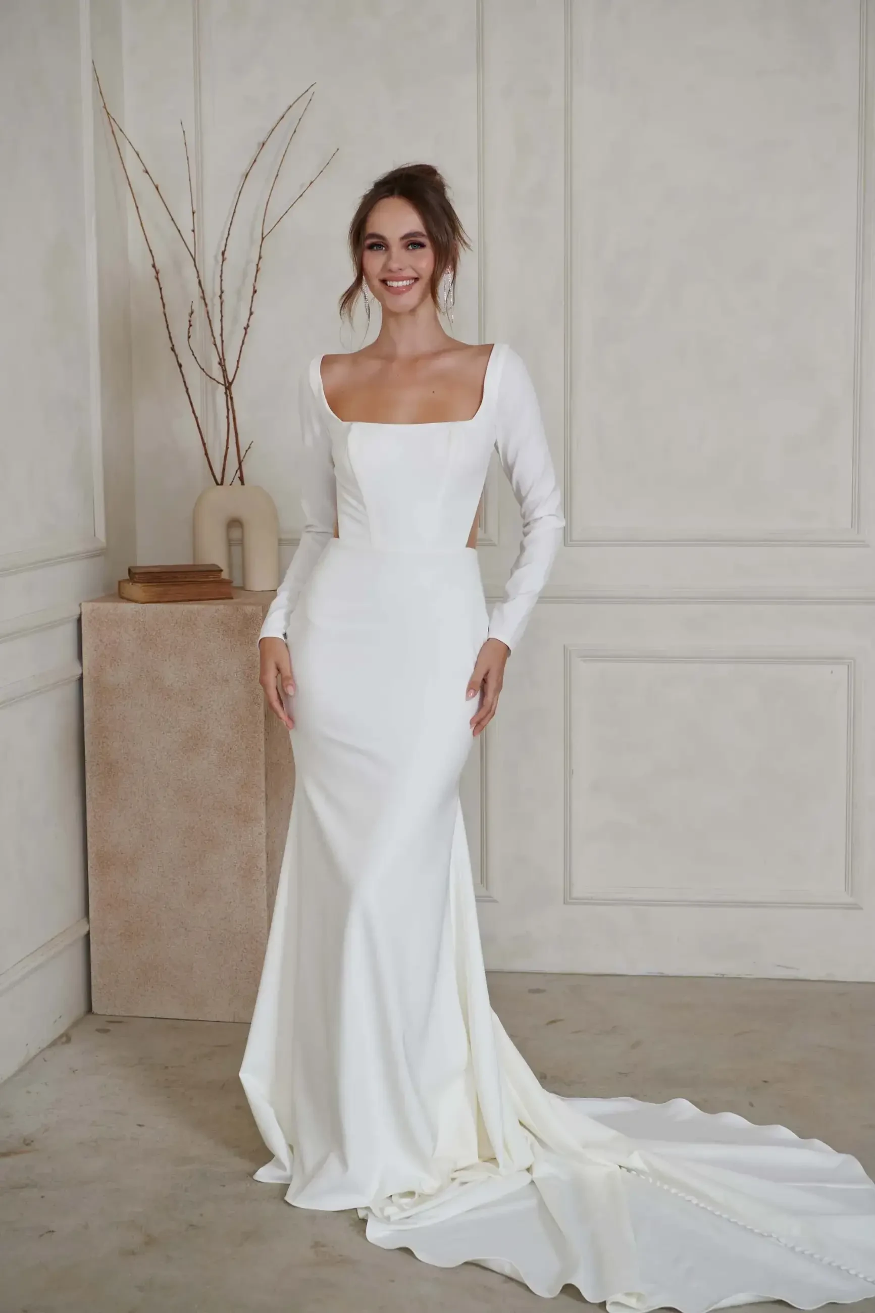 A woman in a fitted long-sleeve, white wedding gown with a square neckline stands smiling in a minimalistic room. The dress features a long train.