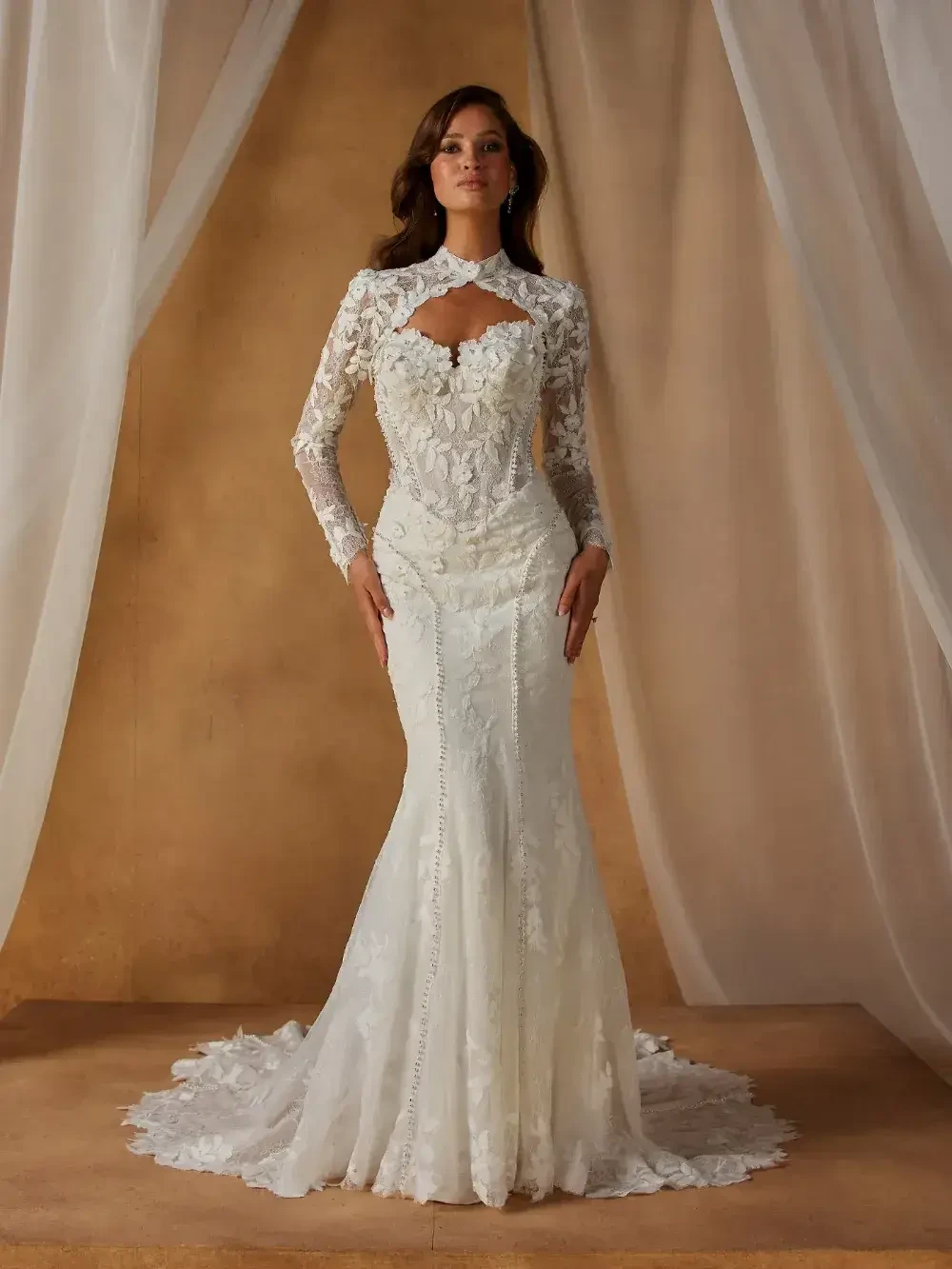 A woman in a detailed lace wedding gown stands confidently. The dress features long sleeves and a keyhole neckline, set against elegant, flowing curtains.