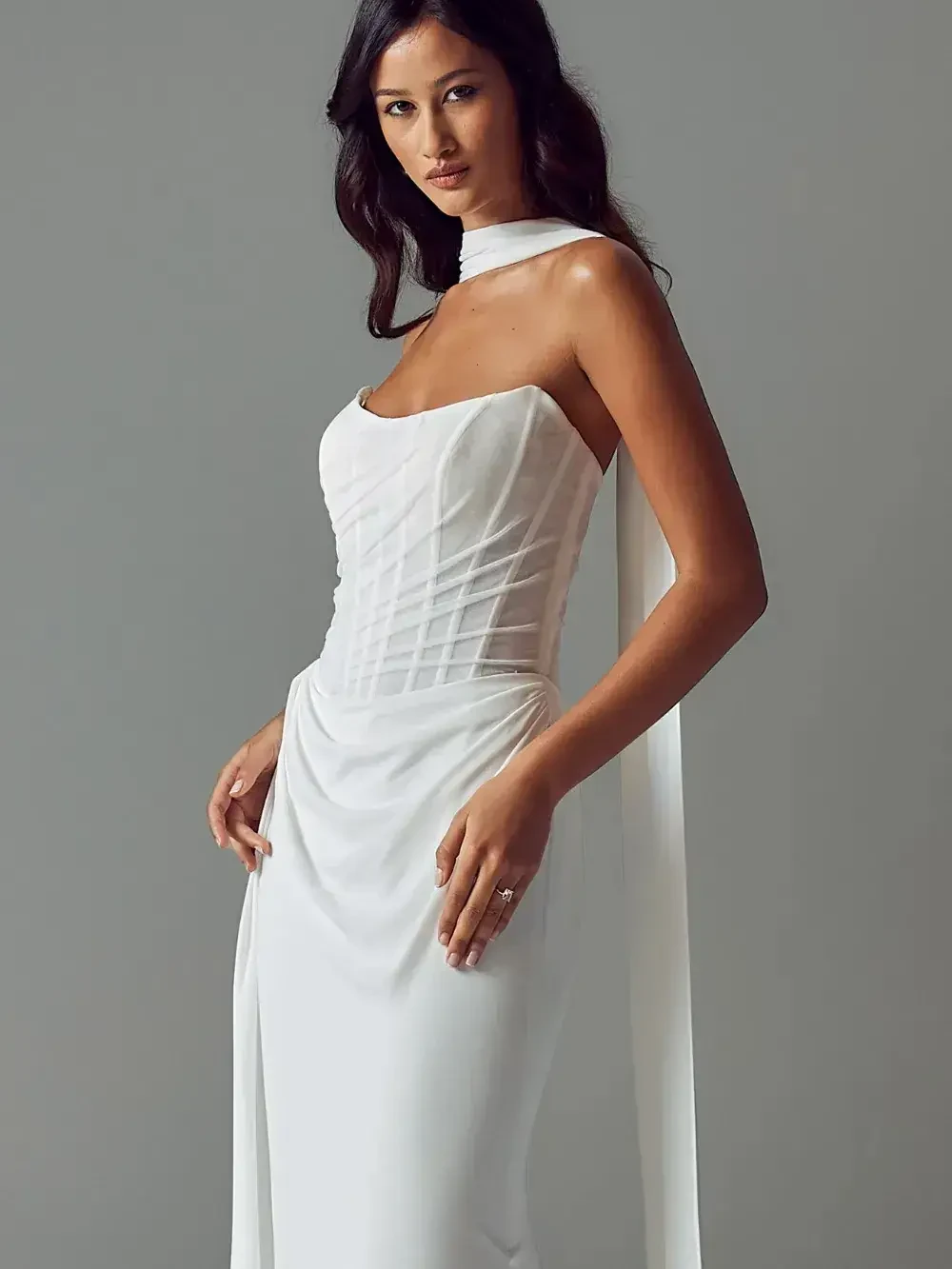 Woman in elegant white strapless gown with minimalist design, featuring a fitted bodice and flowing skirt. She poses confidently against a gray background.