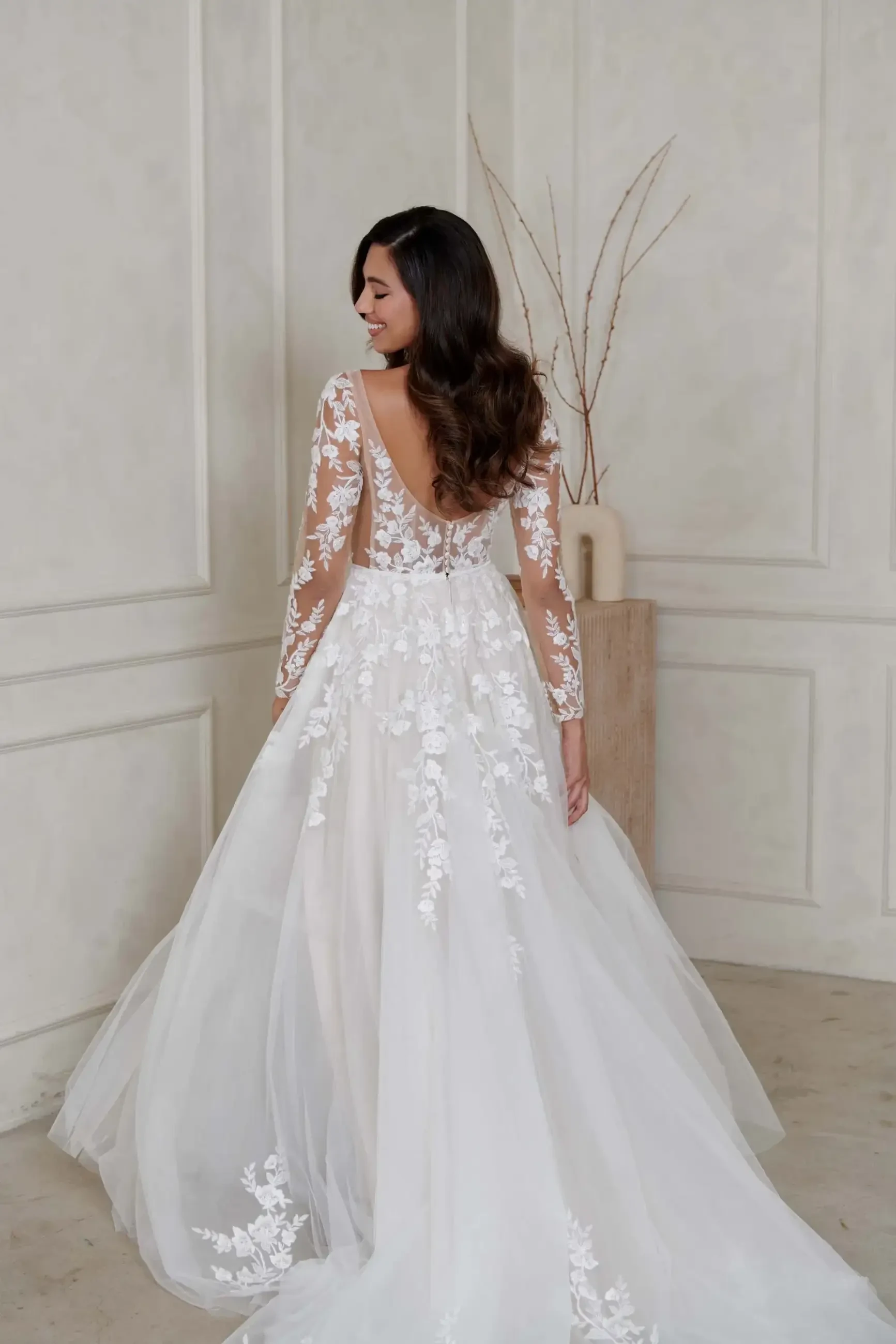 A woman in a lace wedding gown with floral embroidery and long sleeves stands smiling in a simple room with beige walls, creating an elegant and joyful scene.