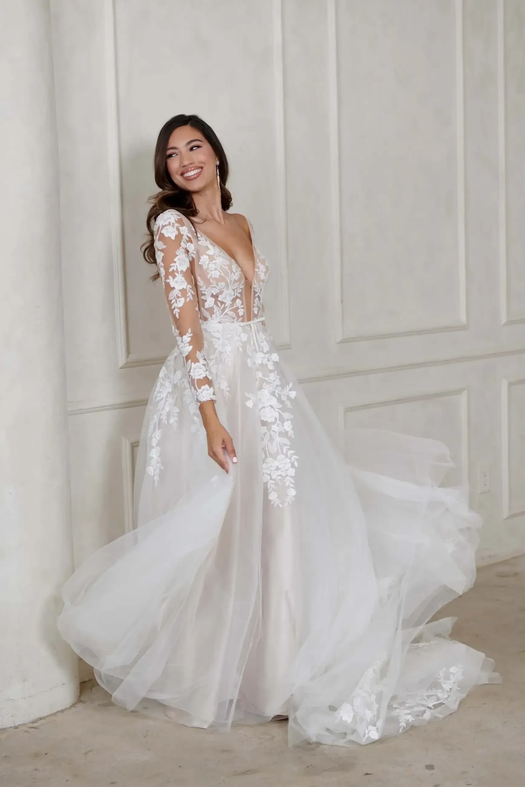 Smiling woman in a flowing white bridal gown with lace details stands in a bright room. She exudes joy and elegance against a neutral backdrop.