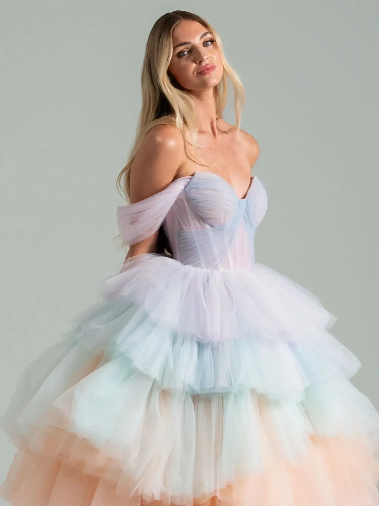 A woman wearing a pastel-colored tulle gown with layered ruffles, looking confidently at the camera.