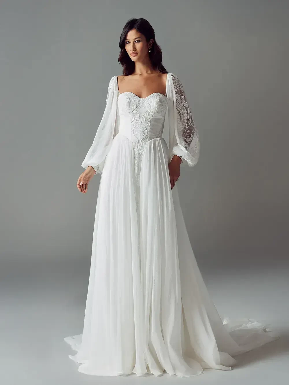 A woman wearing a flowing white gown with long, sheer sleeves and intricate detailing on the bodice stands gracefully against a neutral background.