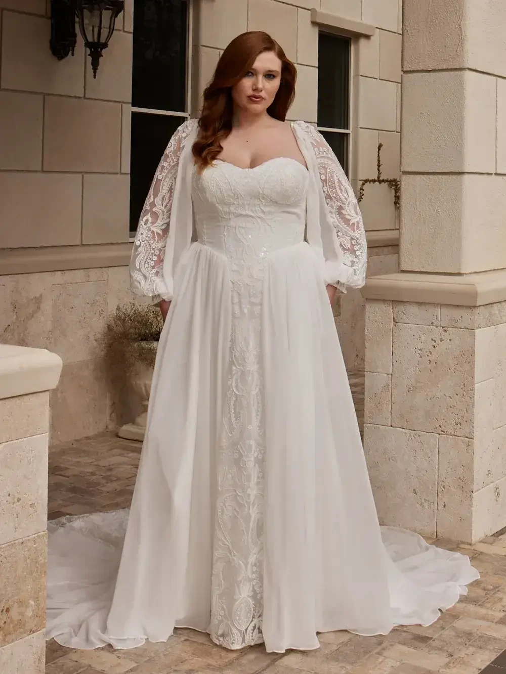 A model wearing a white wedding gown with a heart-shaped neckline and a sheer cape standing on a stone patio.