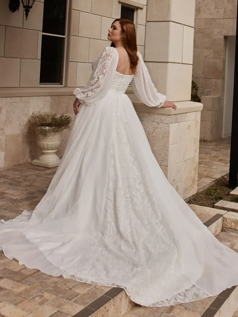 A woman in a white wedding dress with long, flowing sleeves is standing outside, facing away from the camera. The dress features intricate lace detailing and a long train. The backdrop includes a stone wall and a decorative planter.