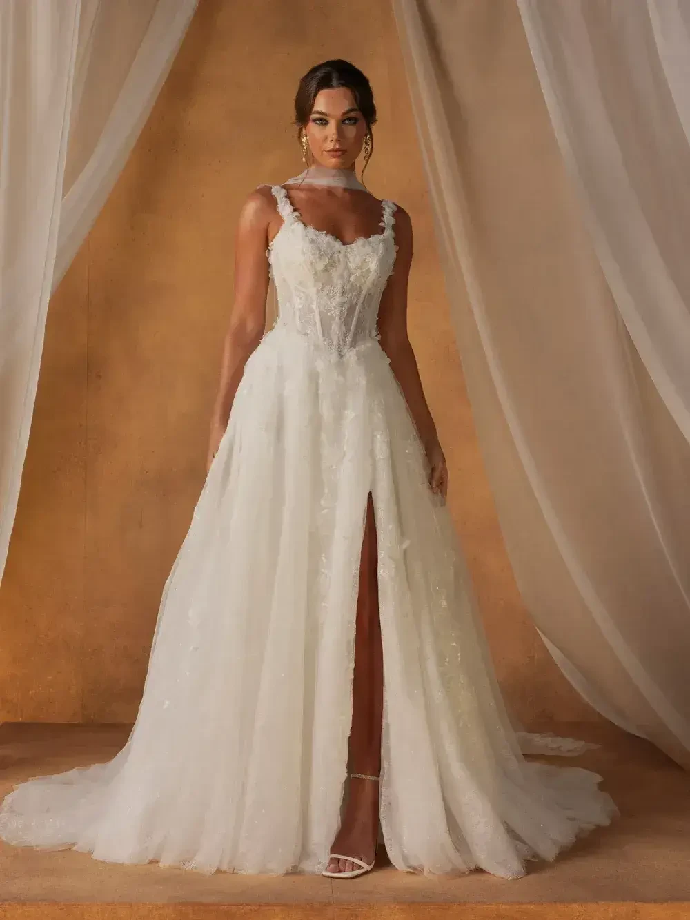 A woman wearing a white wedding dress with a slit, standing on a wooden surface in front of a textured backdrop.
