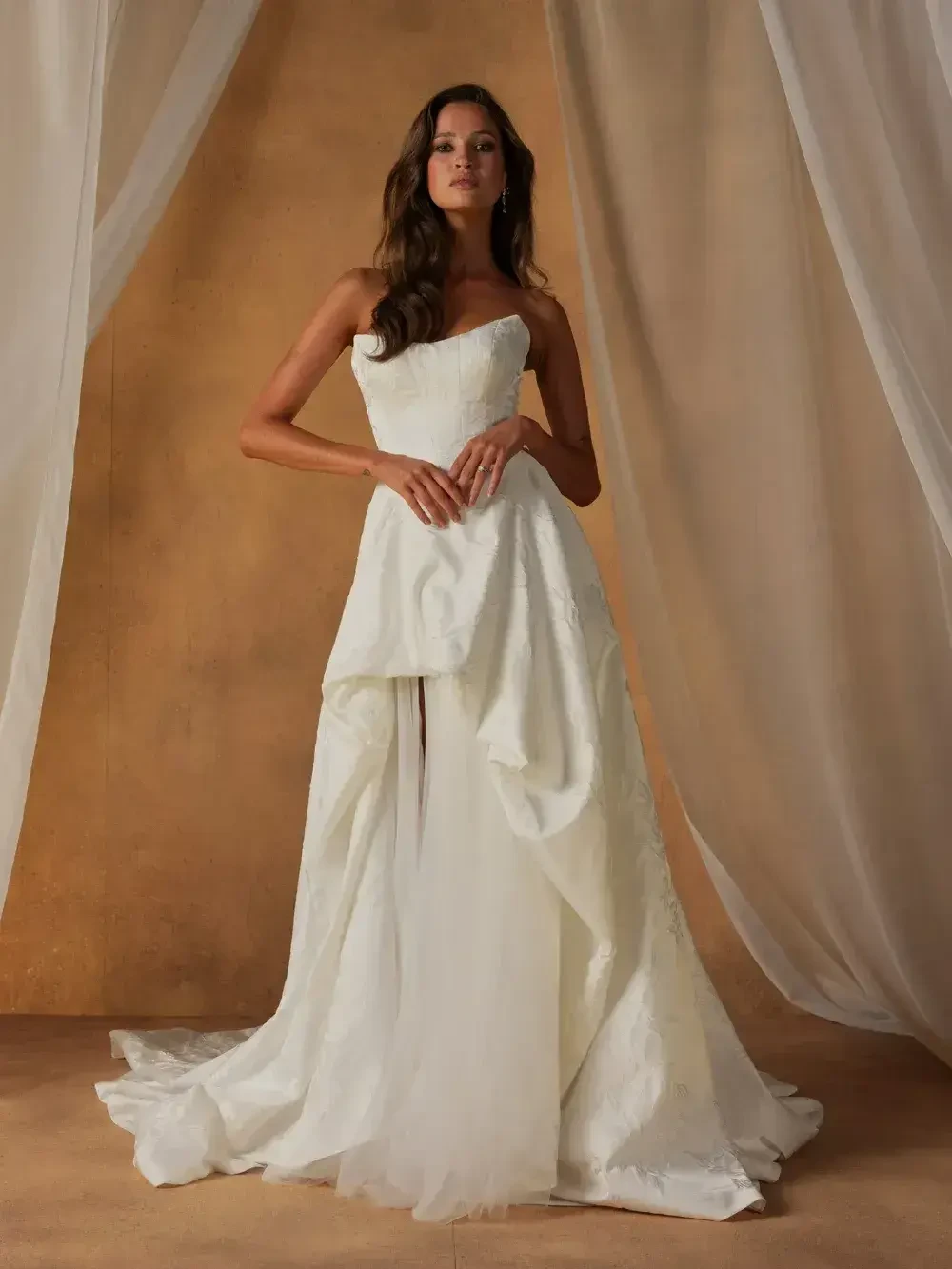 A woman stands elegantly wearing a strapless white wedding dress, featuring a draped design. The background consists of soft fabric drapery, creating a dreamy atmosphere.