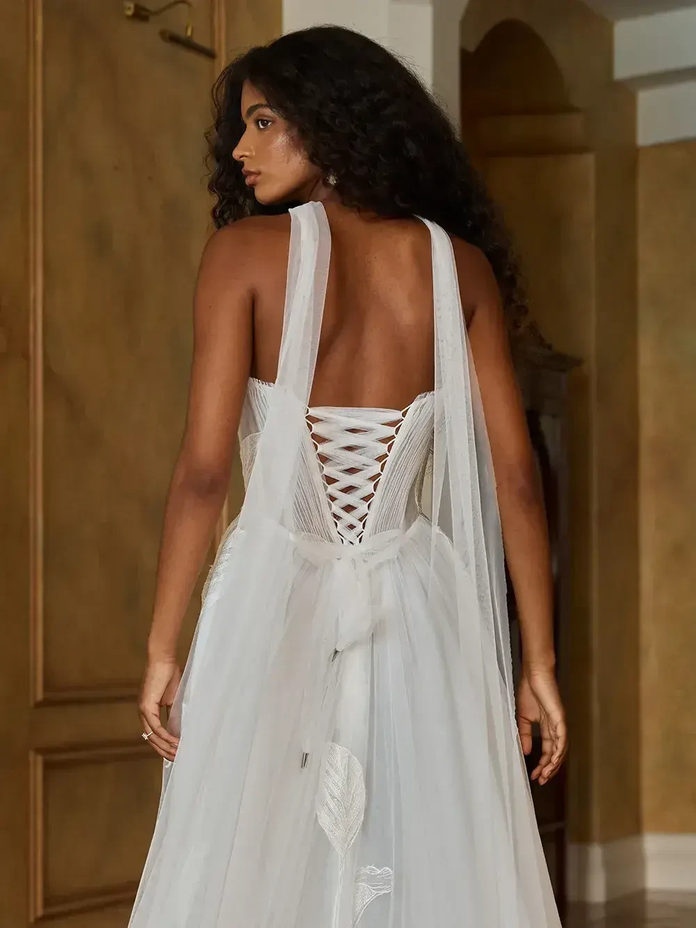 A woman wearing a white gown with a lace-up back, standing with her back to the camera in a softly lit room.