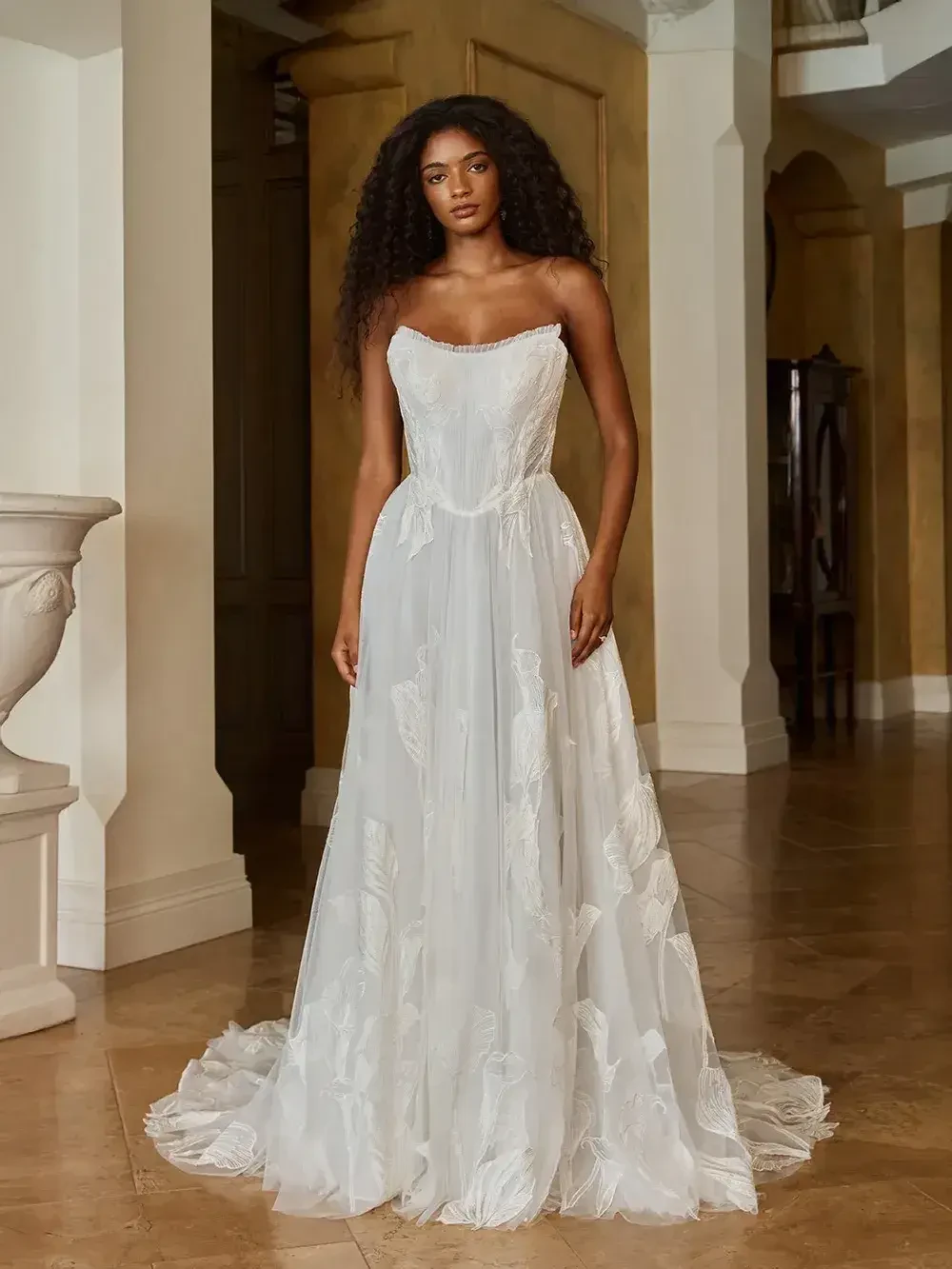 A model wearing a strapless white gown with a flowing skirt, adorned with floral patterns, standing in an elegant indoor setting.