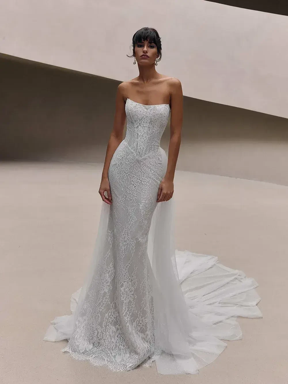 A model wearing a strapless white wedding gown with lace detailing and a flowing train, posing in a minimalist setting.