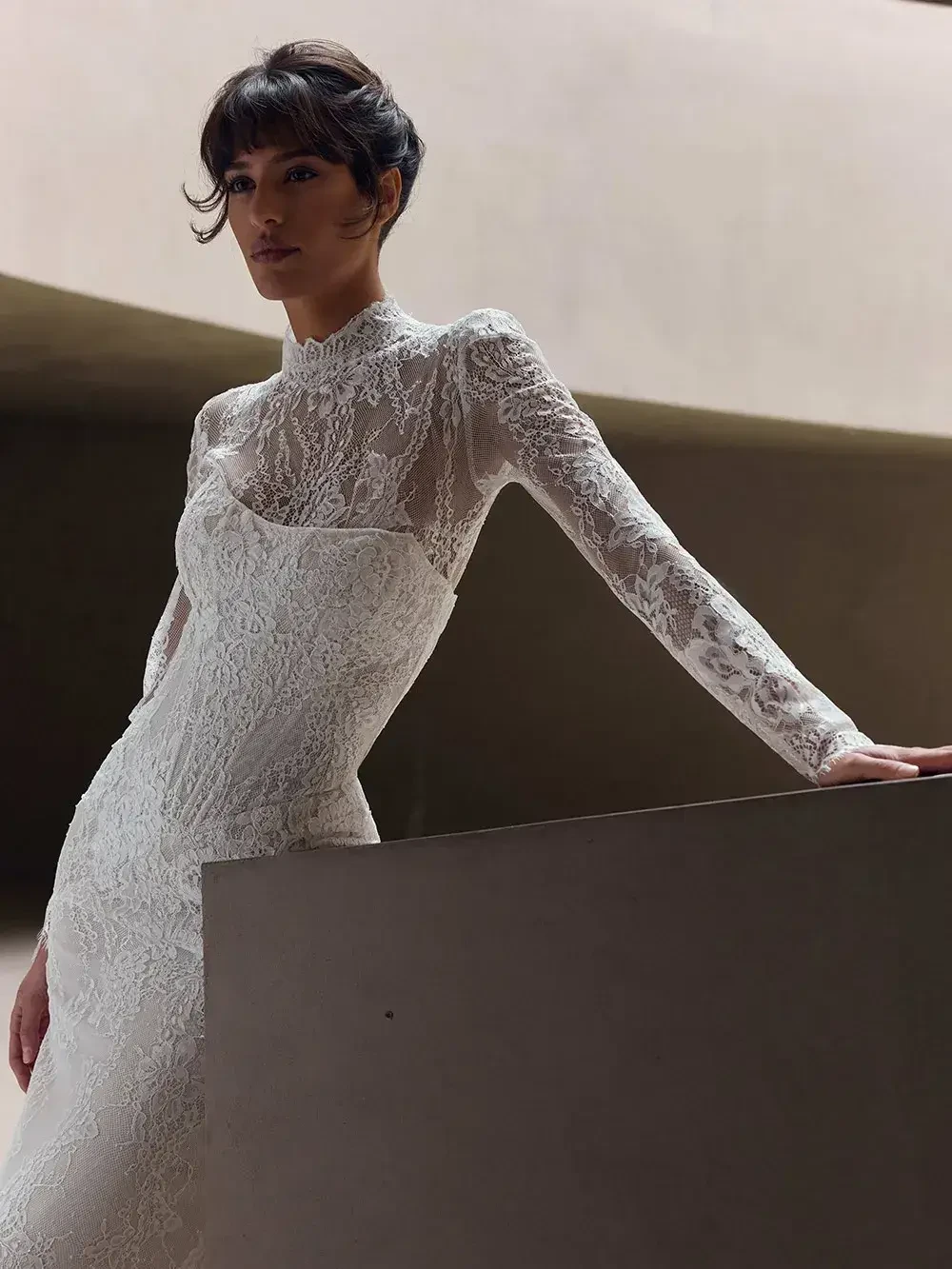 A model poses in an elegant white wedding dress with lace details, featuring long sleeves and a high neckline, set against a minimalist backdrop.