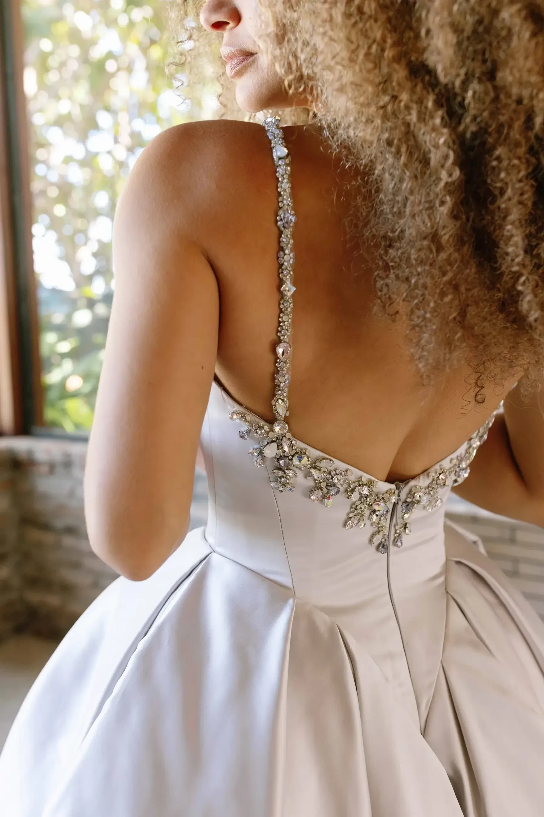 A close-up of the back of a woman wearing a silver dress with intricate beadwork on the straps, showcasing her curly hair and a softly lit background.