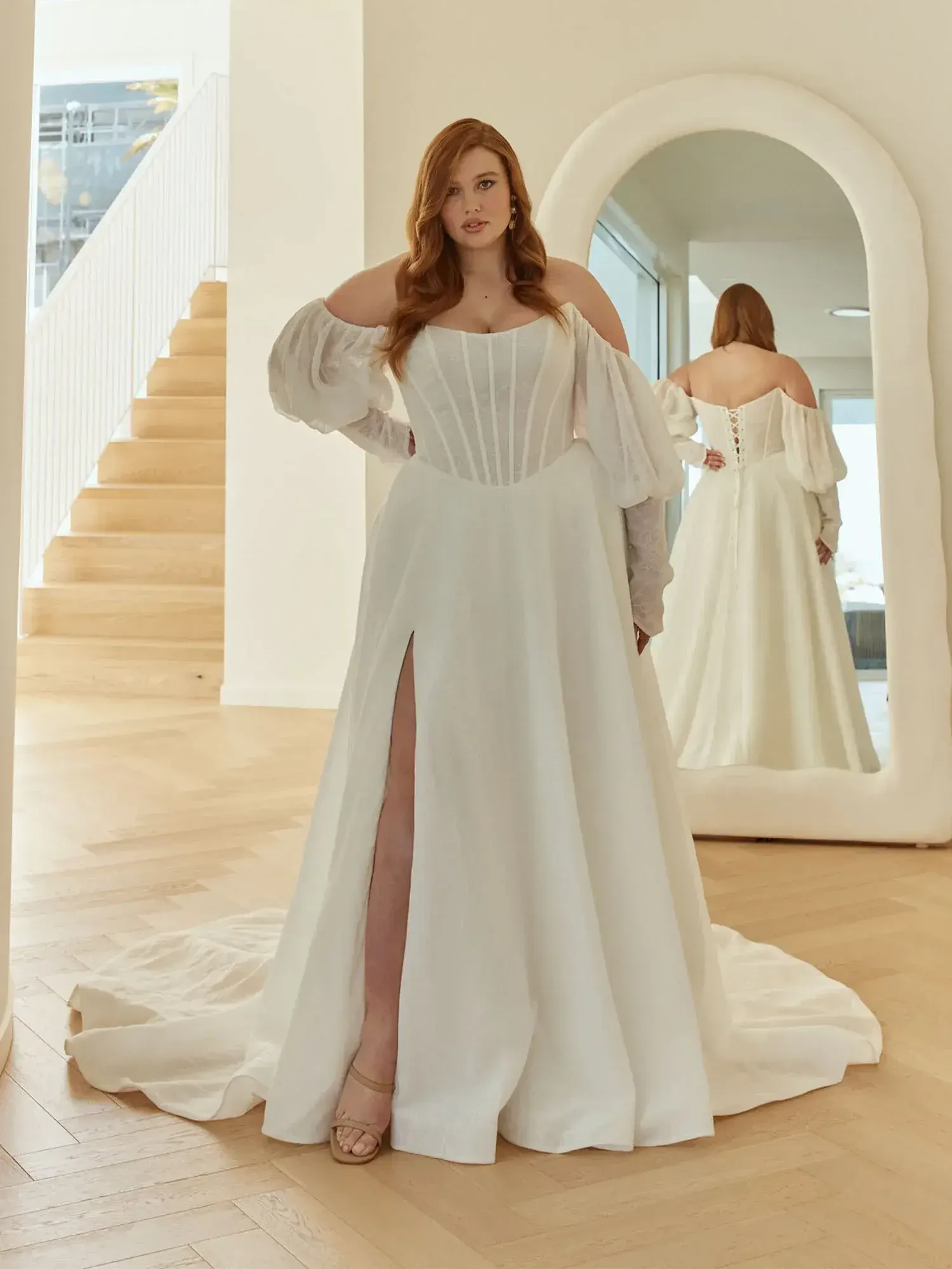 A woman in a white gown stands on a staircase, looking at her reflection in a large mirror. The dress features off-the-shoulder sleeves and a high slit.