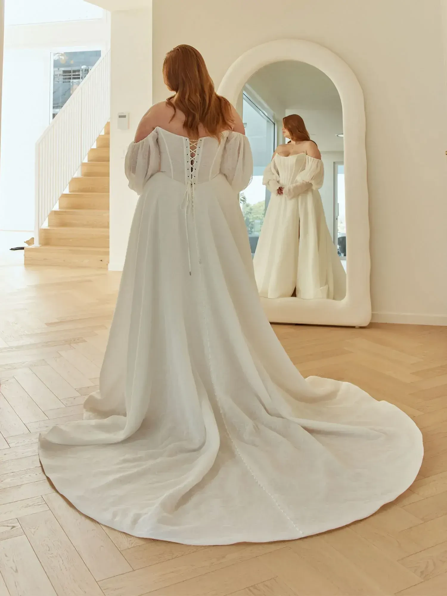 A woman in a white off-shoulder dress with a long train stands in front of a large mirror, viewing her reflection, with a staircase in the background.