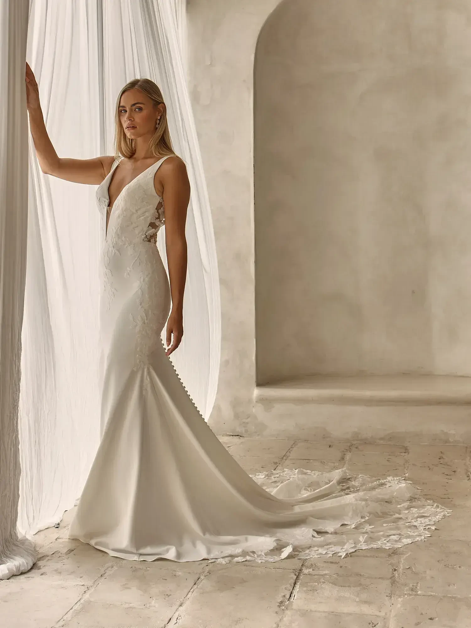 A woman in a sleek, elegant white wedding dress stands beside a flowing curtain in a softly lit room.