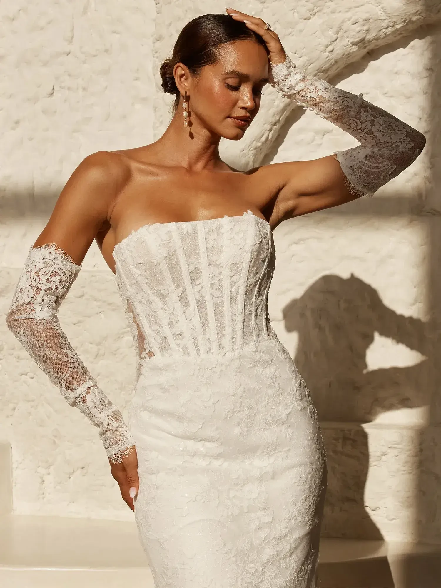 A woman in a strapless white lace wedding dress with long lace sleeves, standing against a textured white wall, with one hand on her head and a serene expression.
