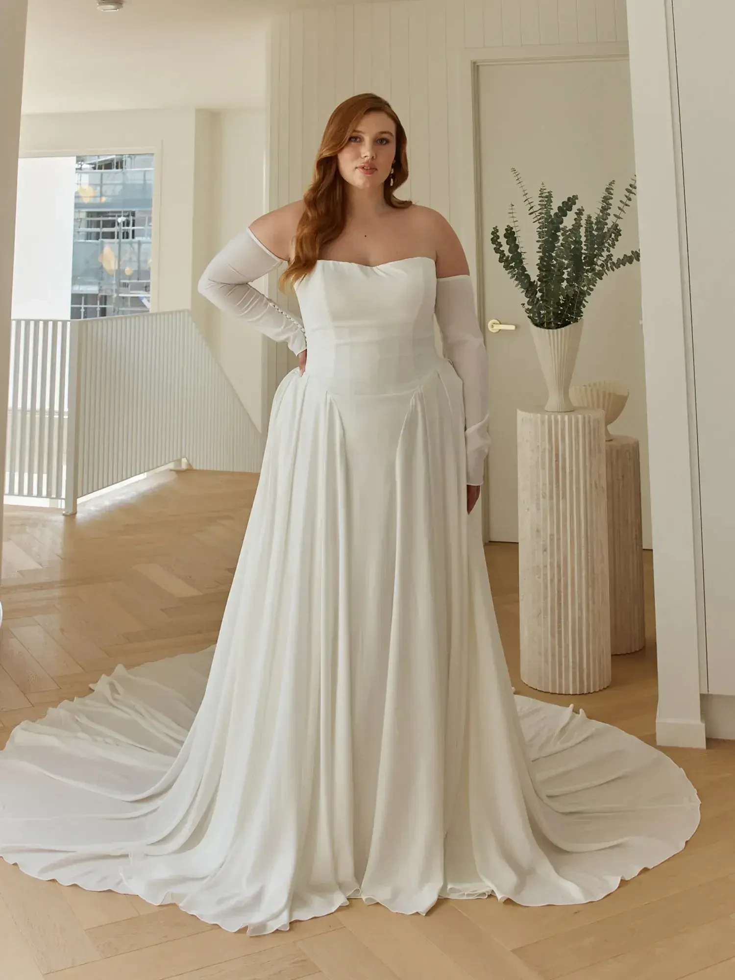 A woman wearing a strapless white wedding dress with long sleeves, standing in a bright, modern room with wooden floors and plants.