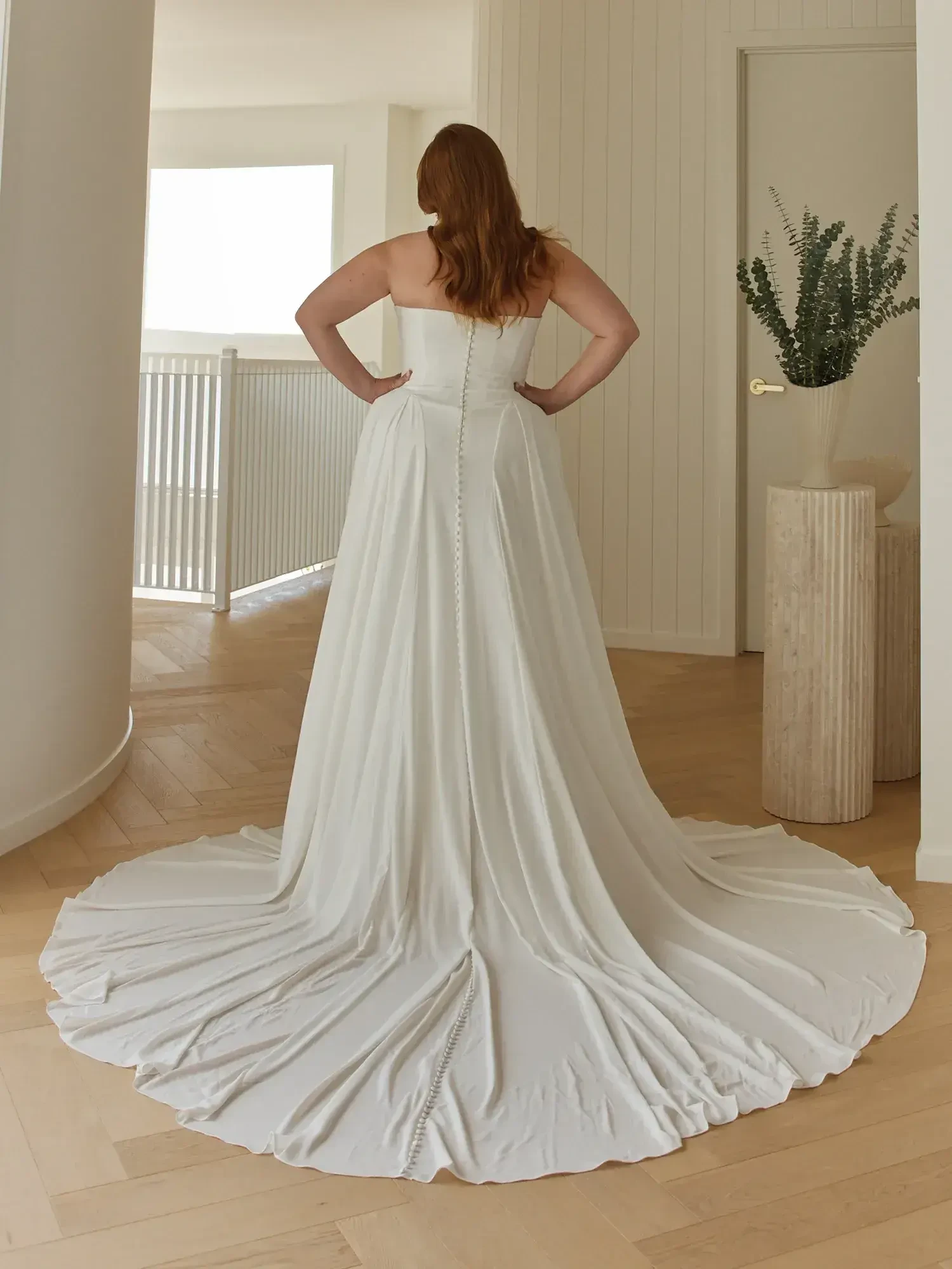 A woman in a white wedding dress, standing with her back to the camera, showcasing the train of the dress as she poses in a bright, modern interior.