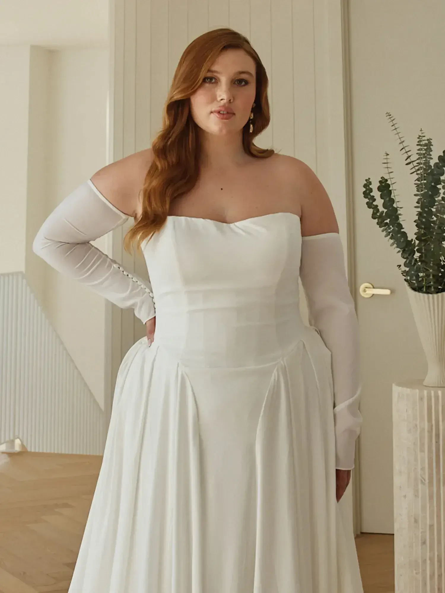 A woman wearing a white off-the-shoulder dress stands in a bright room with modern decor, featuring a soft hairstyle and elegant earrings.